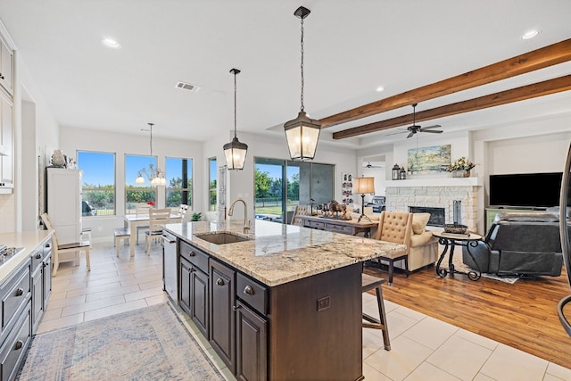 kitchen with sink, beamed ceiling, light hardwood / wood-style floors, a stone fireplace, and an island with sink