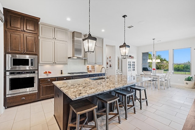 kitchen featuring a kitchen island with sink, wall chimney range hood, sink, appliances with stainless steel finishes, and light stone counters