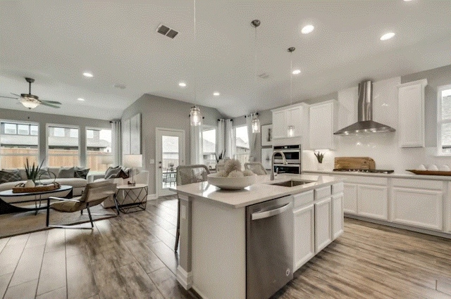 kitchen featuring a kitchen island with sink, stainless steel appliances, light hardwood / wood-style floors, wall chimney range hood, and white cabinetry