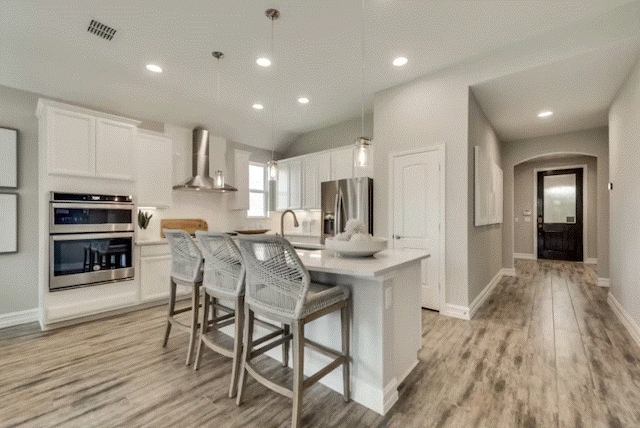 kitchen featuring wall chimney range hood, a center island with sink, hanging light fixtures, appliances with stainless steel finishes, and white cabinetry