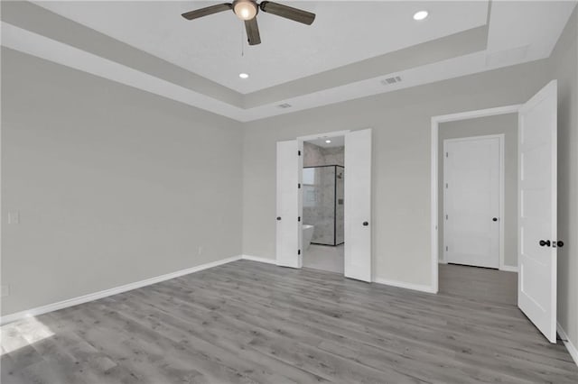 unfurnished bedroom featuring ceiling fan, ensuite bath, a tray ceiling, and hardwood / wood-style floors