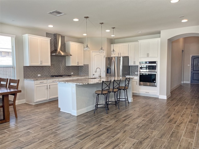 kitchen with appliances with stainless steel finishes, wall chimney exhaust hood, dark hardwood / wood-style flooring, and an island with sink