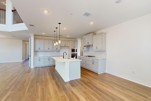 kitchen featuring decorative backsplash, sink, a center island with sink, light hardwood / wood-style floors, and hanging light fixtures