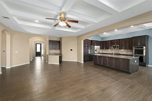 kitchen featuring a kitchen island with sink, appliances with stainless steel finishes, dark brown cabinets, beam ceiling, and backsplash