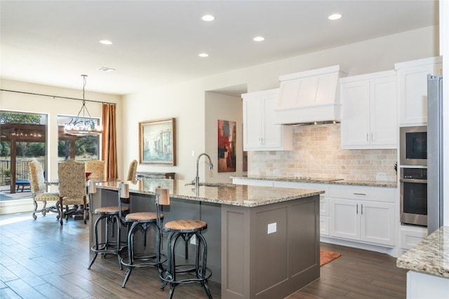 kitchen featuring a kitchen island with sink, custom range hood, pendant lighting, stainless steel oven, and white cabinetry