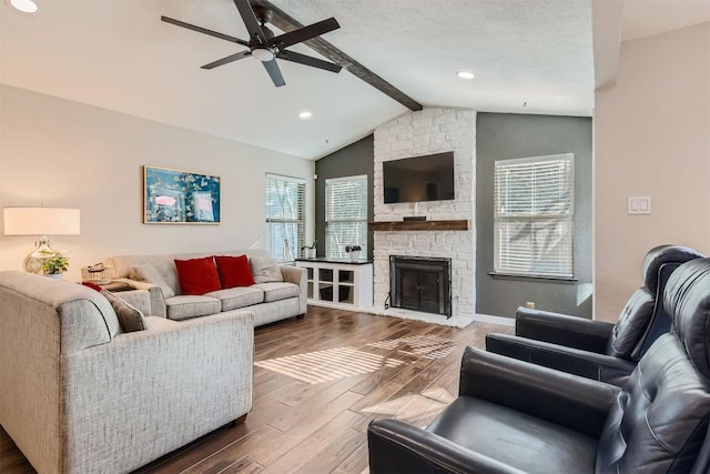 living room with lofted ceiling with beams, ceiling fan, a stone fireplace, a textured ceiling, and wood finished floors