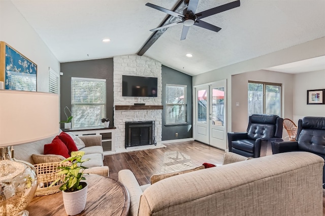 living area with lofted ceiling with beams, a wealth of natural light, wood finished floors, and a stone fireplace