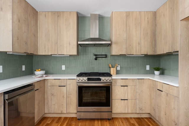 kitchen featuring appliances with stainless steel finishes, light brown cabinetry, wall chimney exhaust hood, and light hardwood / wood-style floors