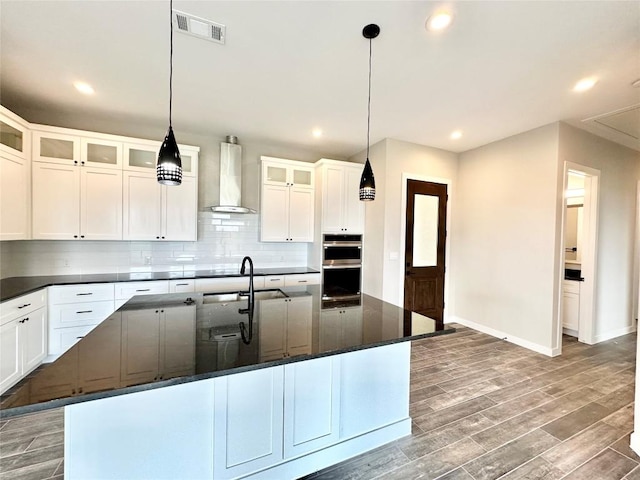 kitchen featuring white cabinetry, hanging light fixtures, wall chimney range hood, dark stone counters, and a center island with sink