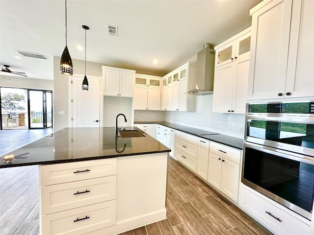 kitchen featuring a kitchen island with sink, wall chimney range hood, sink, double oven, and decorative light fixtures