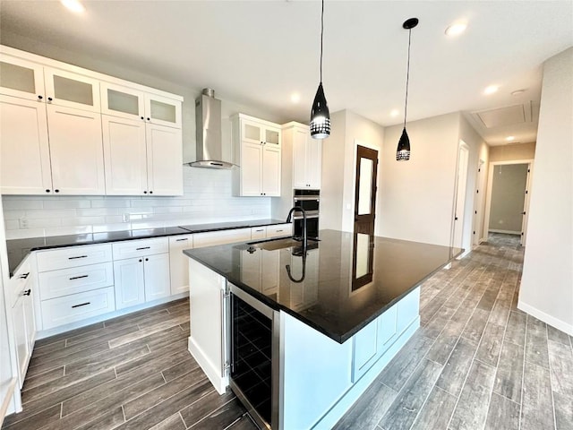 kitchen featuring a kitchen island with sink, wall chimney range hood, sink, white cabinetry, and beverage cooler