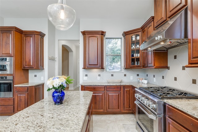 kitchen featuring hanging light fixtures, light stone counters, backsplash, exhaust hood, and appliances with stainless steel finishes