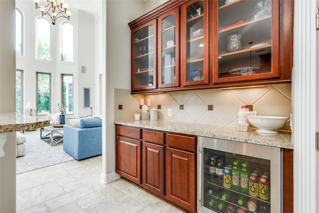 bar with tasteful backsplash, wine cooler, light stone countertops, and an inviting chandelier