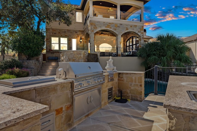 patio terrace at dusk with grilling area, ceiling fan, a balcony, and exterior kitchen