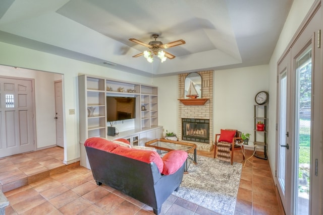 living room with ceiling fan, a tray ceiling, and a brick fireplace