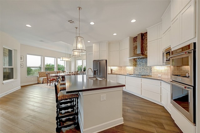 kitchen with a center island with sink, wall chimney exhaust hood, white cabinetry, appliances with stainless steel finishes, and hardwood / wood-style floors