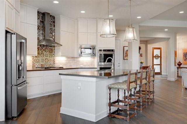 kitchen featuring dark hardwood / wood-style floors, white cabinets, wall chimney range hood, appliances with stainless steel finishes, and a center island with sink