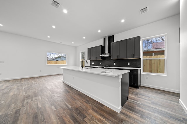 kitchen featuring wall chimney range hood, sink, dark wood-type flooring, a kitchen island with sink, and decorative backsplash