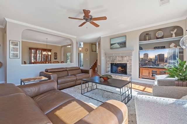 living room with built in shelves, ornamental molding, a stone fireplace, ceiling fan with notable chandelier, and light hardwood / wood-style floors