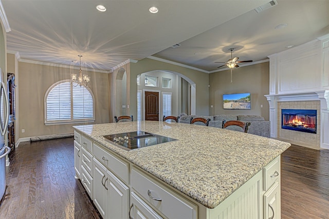 kitchen featuring dark wood-type flooring, ornamental molding, black electric cooktop, an island with sink, and a tiled fireplace