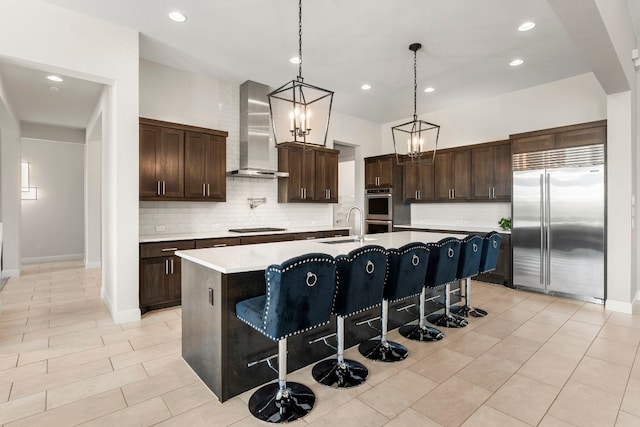 kitchen featuring appliances with stainless steel finishes, a kitchen bar, hanging light fixtures, wall chimney exhaust hood, and a kitchen island with sink