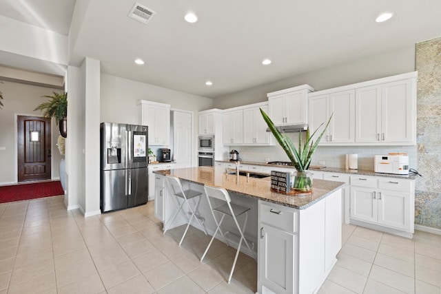 kitchen with light tile patterned floors, light stone counters, a kitchen island with sink, white cabinets, and appliances with stainless steel finishes