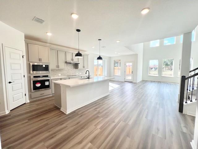 kitchen featuring stainless steel appliances, tasteful backsplash, wood-type flooring, decorative light fixtures, and a kitchen island with sink