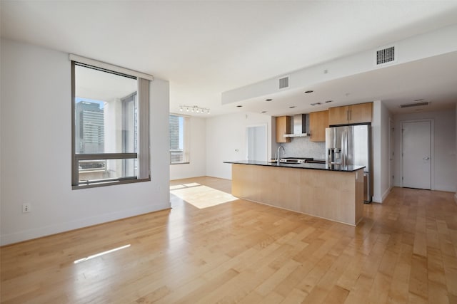 kitchen featuring an island with sink, sink, wall chimney range hood, light hardwood / wood-style floors, and decorative backsplash