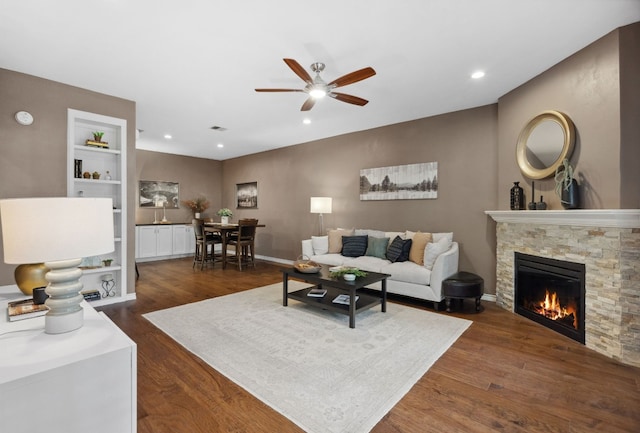 living room with built in features, ceiling fan, dark hardwood / wood-style flooring, and a stone fireplace