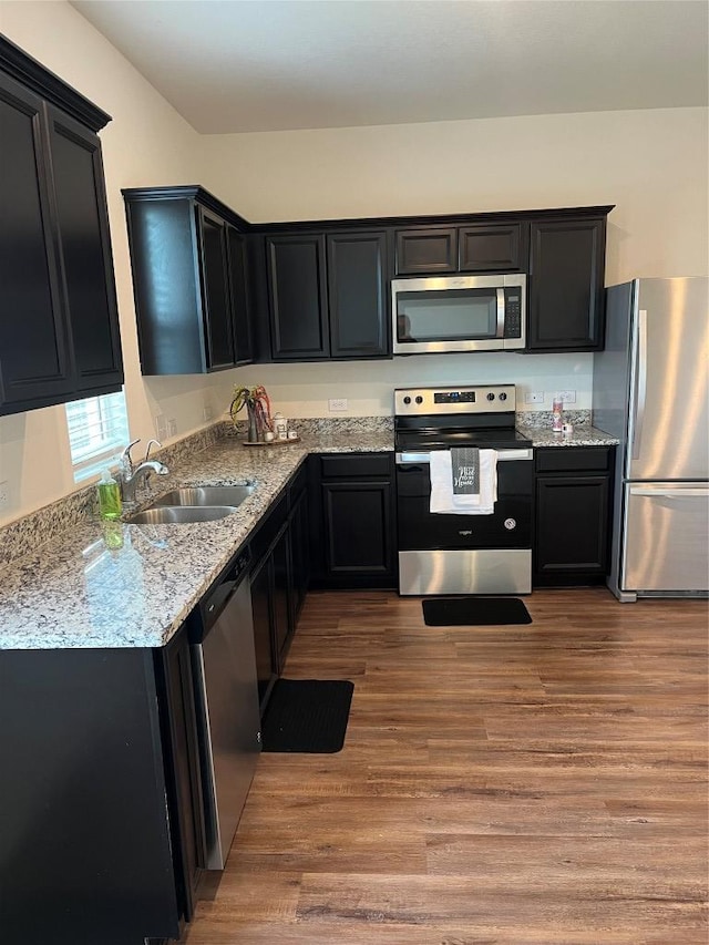 kitchen featuring wood finished floors, appliances with stainless steel finishes, a sink, light stone countertops, and dark cabinetry