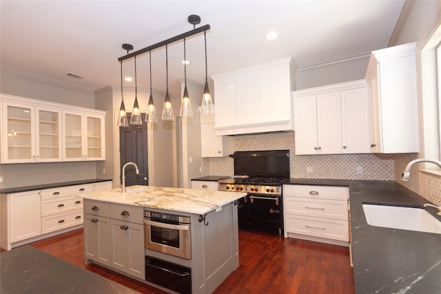 kitchen with a kitchen island with sink, stainless steel oven, sink, hanging light fixtures, and white cabinetry