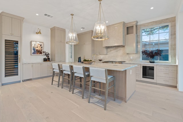 kitchen featuring premium range hood, decorative light fixtures, ornamental molding, an island with sink, and beverage cooler