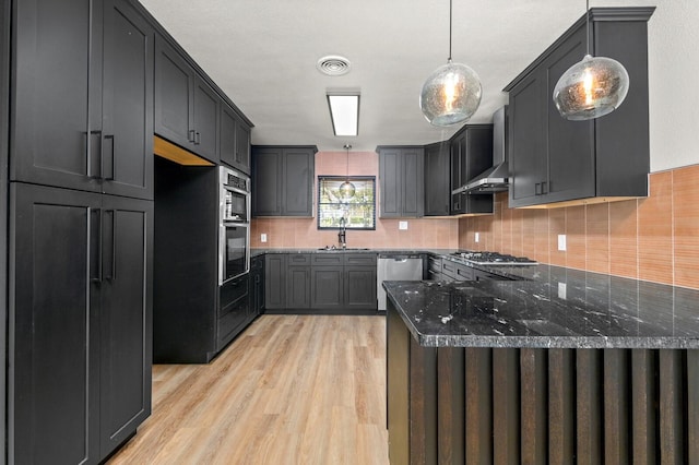 kitchen with wall chimney range hood, sink, hanging light fixtures, appliances with stainless steel finishes, and dark stone counters