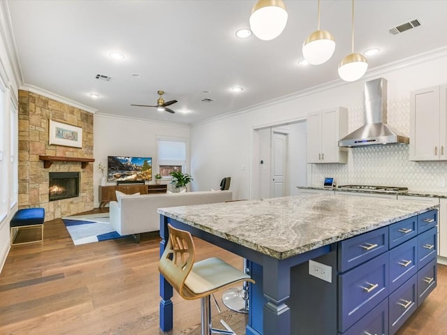 kitchen with ceiling fan, wall chimney range hood, decorative light fixtures, white cabinets, and a kitchen island