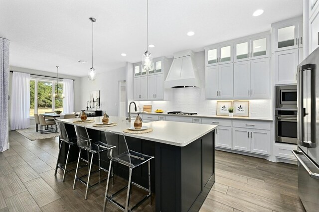 kitchen featuring a kitchen island with sink, custom range hood, white cabinets, decorative light fixtures, and appliances with stainless steel finishes