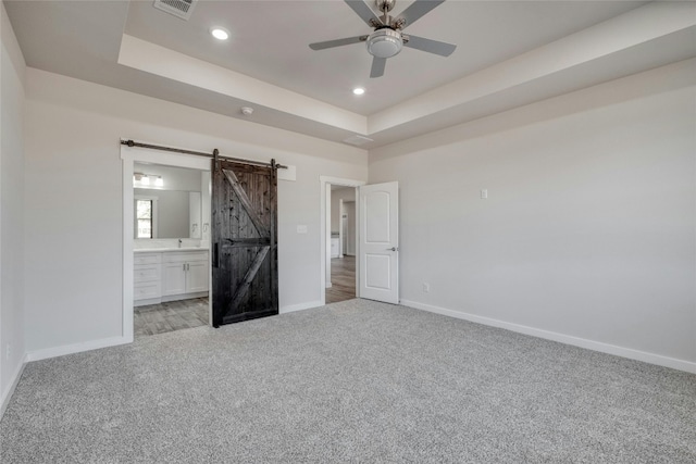 unfurnished bedroom featuring ceiling fan, ensuite bath, a tray ceiling, and a barn door