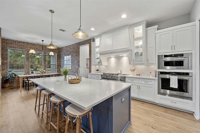 kitchen featuring an island with sink, brick wall, light stone countertops, decorative light fixtures, and white cabinets