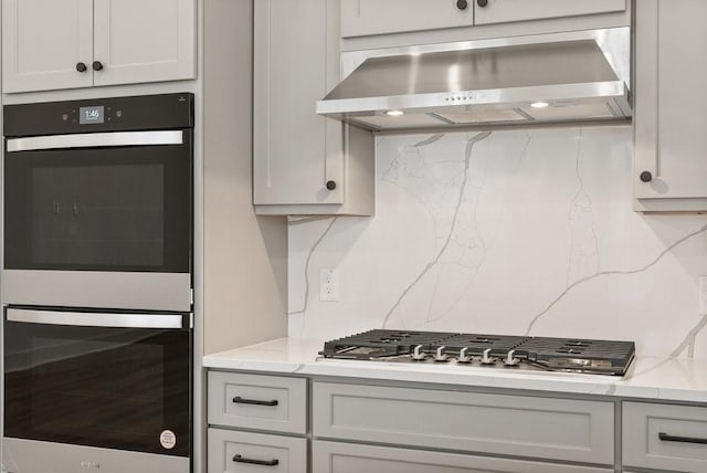 kitchen with wall chimney range hood, double wall oven, light stone countertops, stainless steel gas cooktop, and white cabinets