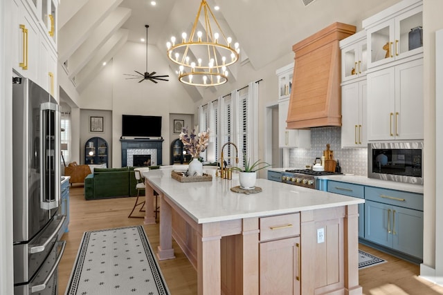 kitchen with appliances with stainless steel finishes, light wood-type flooring, a stone fireplace, white cabinets, and a kitchen island with sink