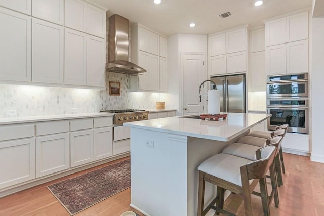 kitchen with wall chimney range hood, appliances with stainless steel finishes, light wood-type flooring, and an island with sink