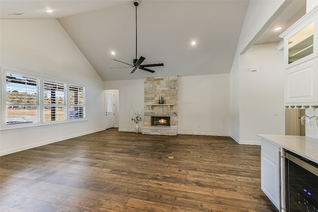 unfurnished living room featuring a stone fireplace, dark hardwood / wood-style flooring, ceiling fan, wine cooler, and high vaulted ceiling