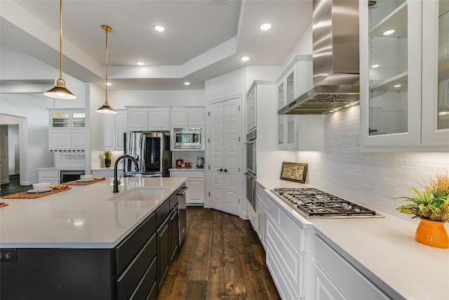 kitchen with wall chimney exhaust hood, sink, hanging light fixtures, appliances with stainless steel finishes, and an island with sink