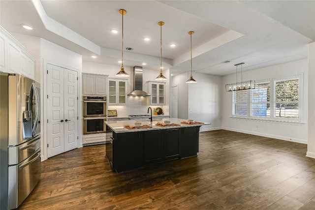 kitchen featuring wall chimney exhaust hood, appliances with stainless steel finishes, white cabinets, and an island with sink