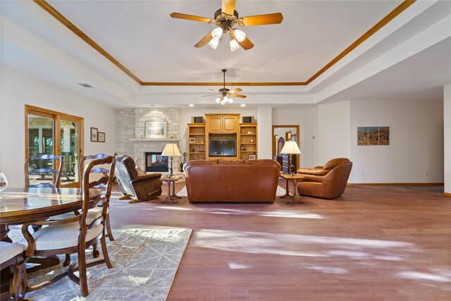 living room featuring hardwood / wood-style floors, a stone fireplace, a raised ceiling, ceiling fan, and crown molding