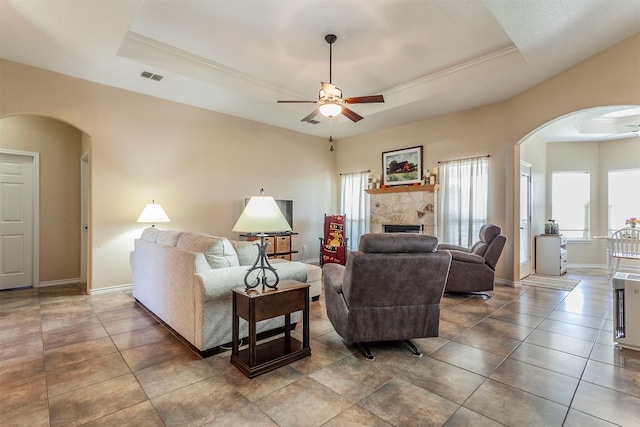 tiled living room with a stone fireplace, ornamental molding, a tray ceiling, and ceiling fan