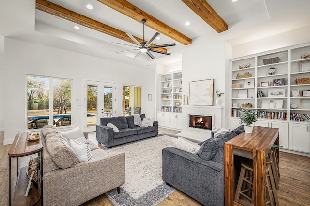 living room featuring a ceiling fan, built in features, beamed ceiling, a warm lit fireplace, and recessed lighting