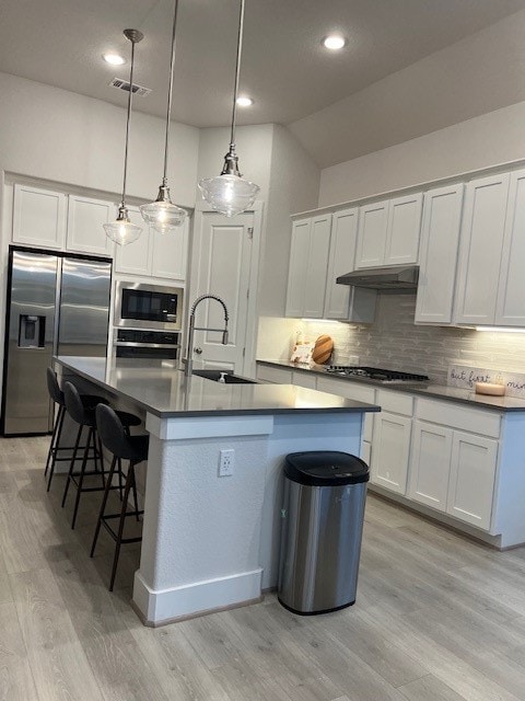 kitchen with white cabinets, a center island with sink, appliances with stainless steel finishes, light wood-type flooring, and decorative light fixtures