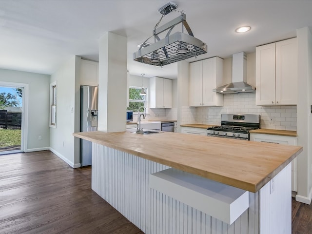 kitchen featuring wall chimney range hood, butcher block countertops, stainless steel appliances, white cabinets, and a center island with sink