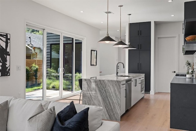 kitchen with pendant lighting, light hardwood / wood-style flooring, stainless steel dishwasher, an island with sink, and light stone counters