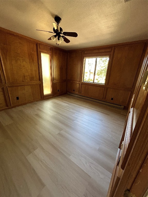 spare room featuring wooden walls, a baseboard heating unit, light wood-type flooring, a textured ceiling, and ceiling fan